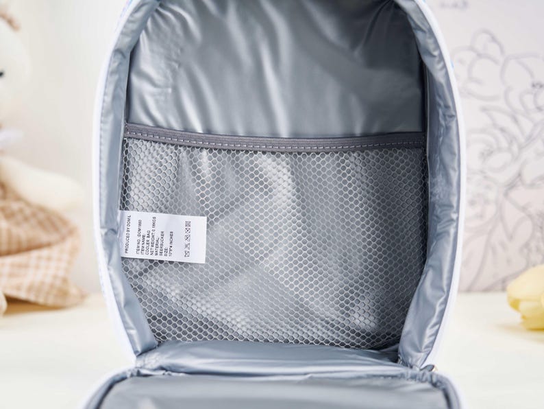 Puede incluir: Vista abierta de una bolsa de almuerzo aislada blanca con interior gris. La bolsa tiene un bolsillo de malla y una etiqueta con texto. El interior est&aacute; forrado con un material aislante reflectante.