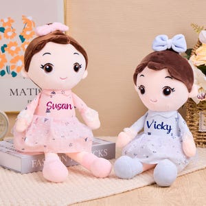 Puede incluir: Dos muñecas de peluche con cabello castaño y lazos, vestidas con vestidos. Una muñeca lleva un vestido rosa con el nombre "Susan", y la otra un vestido azul claro con el nombre "Vicky". Están sentadas sobre una superficie color crema.