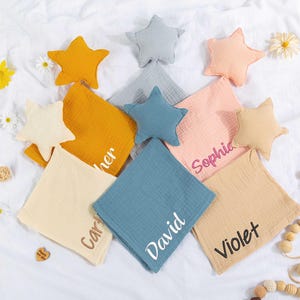 Può includere: Una collezione di articoli per bambini, tra cui peluche a forma di stella e quadrati di mussola personalizzati. I quadrati sono in vari colori, come giallo, blu e rosa, con nomi ricamati. Margherite e giocattoli di legno sono sparsi.