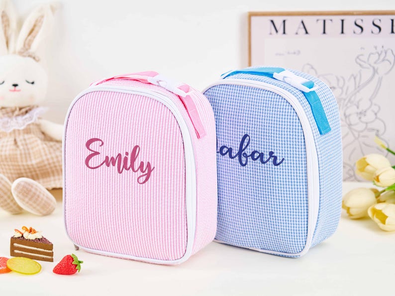 Puede incluir: Dos bolsas de almuerzo personalizadas. Una es rosa con rayas verticales y el nombre "Emily" en cursiva. La otra es azul con un estampado de cuadros y el nombre "Afar" en cursiva. Ambas tienen asa y cremallera.