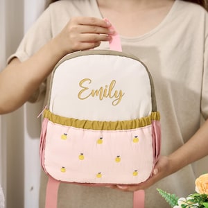 Puede incluir: Una pequeña mochila personalizada en tonos rosa, crema y verde oliva. La mochila presenta el nombre "Emily" bordado en oro. El bolsillo inferior está decorado con fruta amarilla bordada.