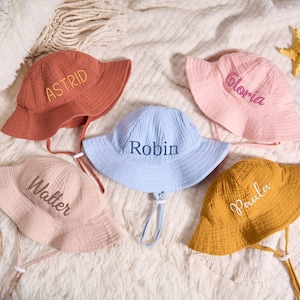 May include: Five personalised bucket hats in various colours, including terracotta, pale pink, baby blue, mustard yellow, and blush pink. Each hat features a name embroidered on it: Astrid, Gloria, Robin, Walter, and Paula. They all have chin straps.