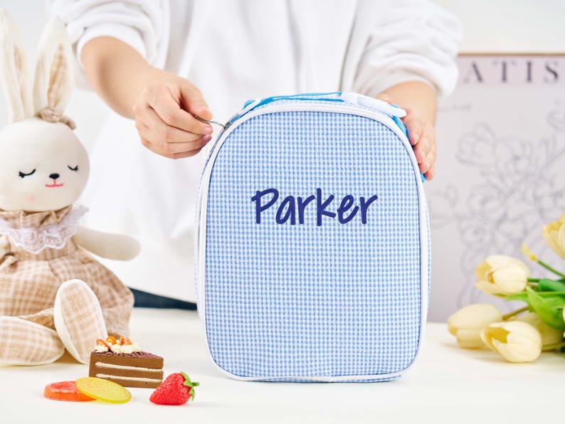 Puede incluir: Una bolsa de almuerzo a cuadros azules y blancos con el nombre "Parker" en escritura azul marino. La bolsa tiene un ribete blanco y un asa azul. Un conejito de peluche, comida de juguete y flores est&aacute;n en el fondo.