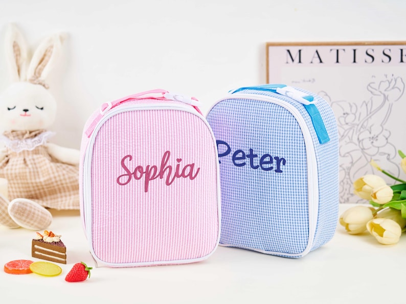 Puede incluir: Dos loncheras personalizadas. Una es rosa con rayas verticales y el nombre "Sophia" en cursiva. La otra es azul a cuadros con el nombre "Peter" en azul. Ambas tienen un asa superior y cremallera blanca.