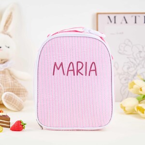 Puede incluir: Bolsa de almuerzo a rayas rosas y blancas con el nombre "MARIA" en fuente manuscrita. La bolsa tiene forma redondeada y cremallera blanca. Un conejito de peluche y objetos decorativos en el fondo.