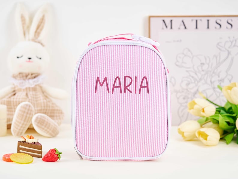 Puede incluir: Bolsa de almuerzo a rayas rosas y blancas con el nombre "MARIA" en fuente manuscrita. La bolsa tiene forma redondeada y cremallera blanca. Un conejito de peluche y objetos decorativos en el fondo.