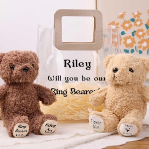May include: Two plush teddy bears, one brown and one tan, sit side-by-side. The brown bear has "Ring Bearer" and "Riley 8-8-25" printed on its feet. The tan bear has "Flower Girl" and "Steve 02-09-25" printed on its feet. A clear bag with "Riley Will you be our Ring Bearer" is in the background.