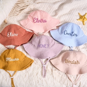 May include: Six pastel-coloured baby sun hats with embroidered names. The hats are in pink, orange, yellow, lavender, and blue. Each hat has a chin strap. The names embroidered are Elvira, Oliver, Maura, Honey, Carter, and Beulah.