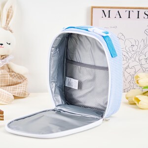Puede incluir: Bolsa de almuerzo abierta a cuadros azules y blancos con interior blanco y bolsillo de malla. La bolsa tiene un asa y cremallera azules. Un conejito de peluche y una impresi&oacute;n de Matisse est&aacute;n en el fondo.