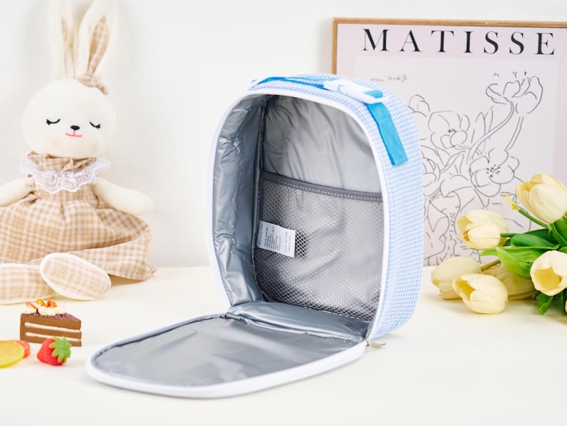 Puede incluir: Bolsa de almuerzo abierta a cuadros azules y blancos con interior blanco y bolsillo de malla. La bolsa tiene un asa y cremallera azules. Un conejito de peluche y una impresi&oacute;n de Matisse est&aacute;n en el fondo.