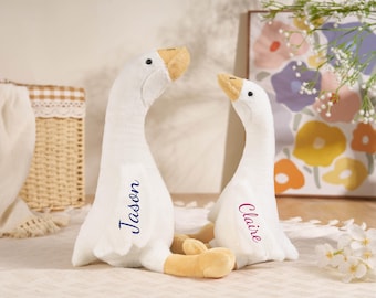 May include: Two plush white goose toys with orange beaks and feet. The larger goose has the name Jason embroidered in blue on its wing, and the smaller goose has the name Claire embroidered in magenta on its wing, both in a cursive script.
