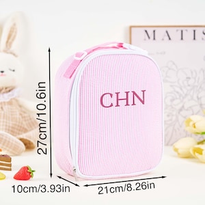 Puede incluir: Bolsa de almuerzo rosa a rayas con las iniciales "CHN". La bolsa mide 27 cm de alto, 21 cm de ancho y 10 cm de profundidad. Tiene un asa y cierre de cremallera. La bolsa est&aacute; junto a un conejito de peluche y comida de juguete.