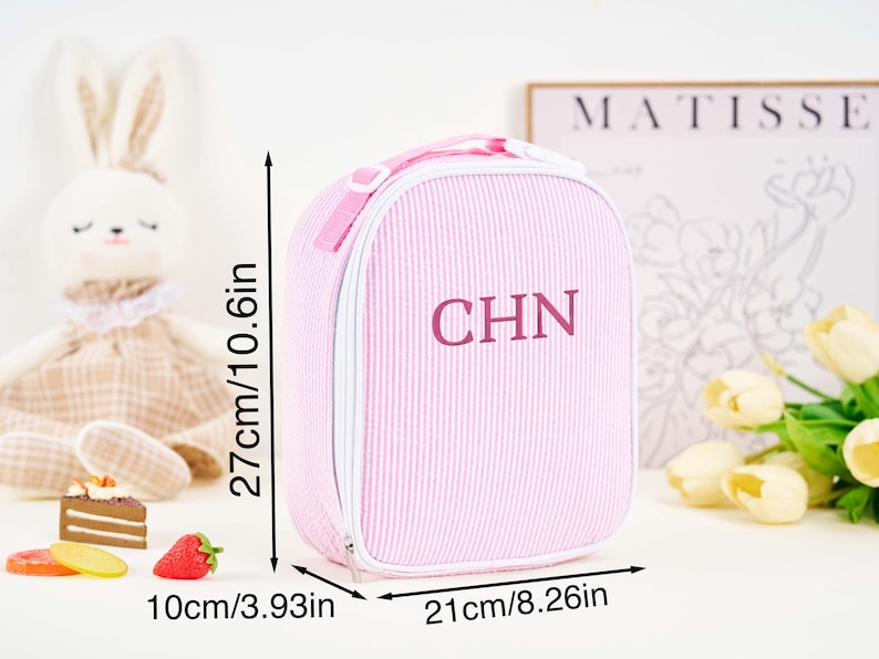 Puede incluir: Bolsa de almuerzo rosa a rayas con las iniciales "CHN". La bolsa mide 27 cm de alto, 21 cm de ancho y 10 cm de profundidad. Tiene un asa y cierre de cremallera. La bolsa est&aacute; junto a un conejito de peluche y comida de juguete.