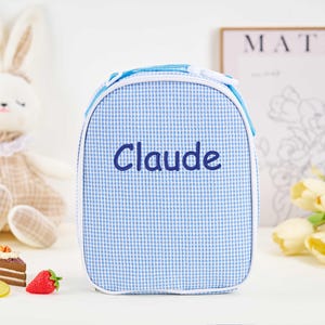 Puede incluir: Una bolsa de almuerzo a cuadros azules y blancos con el nombre "Claude" bordado en azul marino. La bolsa tiene un ribete blanco y una cremallera azul. Un conejito de peluche y una naturaleza muerta de comida est&aacute;n en el fondo.
