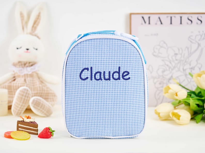 Puede incluir: Una bolsa de almuerzo a cuadros azules y blancos con el nombre "Claude" bordado en azul marino. La bolsa tiene un ribete blanco y una cremallera azul. Un conejito de peluche y una naturaleza muerta de comida est&aacute;n en el fondo.