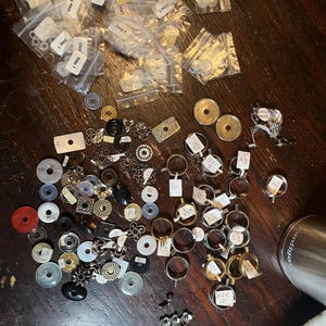 May include: A collection of assorted metal and plastic jewelry components, including rings, circular settings, and small charms. Many items are labeled with white paper tags marked with numbers. Several small clear plastic bags contain small parts labeled Step 1.