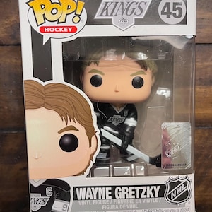 May include: A Wayne Gretzky vinyl figure from the Los Angeles Kings hockey team. The figure is in a Pop! box with the team logo and NHL branding. The figure is wearing a black and white hockey uniform and holding a hockey stick.