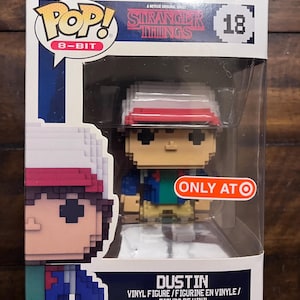 May include: A boxed 8-bit Dustin vinyl figure from the Netflix series Stranger Things. The figure has a red and white hat, and is wearing a blue jacket and tan pants. The box has the text "POP! 8-BIT" and "ONLY AT Target".