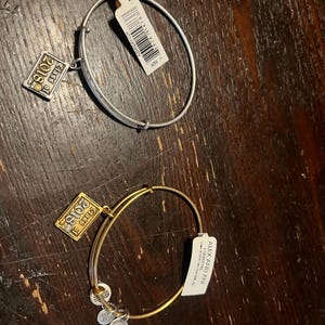 May include: Two Alex and Ani bangles. One is silver-toned, the other gold-toned. Each has a charm that reads "Class of 2018". Both have attached tags. The bangles are on a dark brown wooden surface.