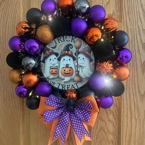May include: A Halloween wreath featuring purple, orange, and black ornaments surrounding a central circular sign with three ghosts and the text Trick or Treat. A large orange and purple polka dot bow with a black spider sits at the bottom.
