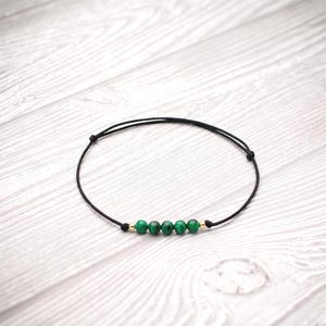 May include: A delicate bracelet featuring a thin black cord with five small, round, green malachite beads and two tiny gold-toned metallic spacers. The bracelet is designed with an adjustable sliding knot mechanism to fit various wrist sizes.
