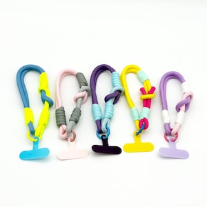 May include: Five colorful phone charms with braided cords in blue, pink, purple, yellow, and lavender. Each charm has a different color combination, with a matching colored tab for phone attachment. The cords are knotted for a decorative touch.