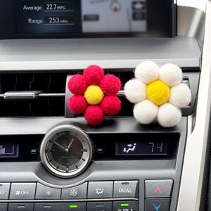 May include: Two flower-shaped car air fresheners. One is red with a yellow center, the other is white with a yellow center. They are placed on a car's dashboard air vent. The car's clock and temperature display are also visible.
