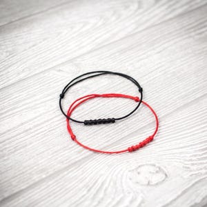 May include: Two thin cord bracelets, one black and one red, each featuring a series of small, uniform knots arranged in a line along the cord. The bracelets are circular and constructed from smooth, flexible material with a simple, minimalist design.