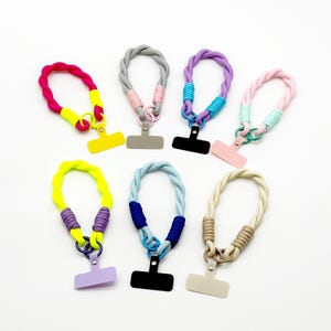 May include: A collection of colorful phone lanyards. Each lanyard features a braided cord in various colors, including pink, yellow, gray, purple, blue, and beige. The lanyards have a small, rectangular phone holder at the bottom.