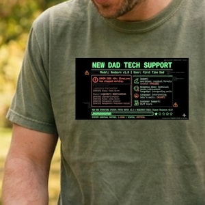 May include: Olive green t-shirt with the text "NEW DAD TECH SUPPORT" in a green and black design. A person is pushing a stroller with a baby wearing a cream-colored beanie. The baby is smiling.