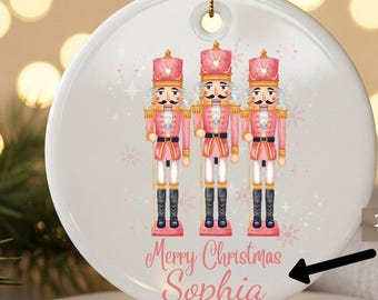 May include: A round Christmas ornament featuring three pink and gold nutcracker soldiers. The ornament has the text "Merry Christmas Sophia" in pink script. A gold ribbon is included for hanging.
