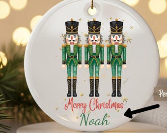 May include: A white ceramic Christmas ornament featuring three toy soldiers in green uniforms and black hats. The ornament has the text "Merry Christmas Noah" in red and green script. A gold ribbon is included.