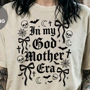 A person wearing a cream-colored t-shirt with black gothic lettering that reads In my God Mother Era and skull, bat, and bow graphics.