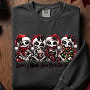 Könnte beinhalten: Ein dunkelgraues Sweatshirt mit Rundhalsausschnitt und einer Grafik von vier Cartoon-Skeletten, die Weihnachtsmützen und festliche Schals tragen. Der Text unter den Skeletten lautet Spooky Moms Have More Festive. Mit geripptem Kragen.