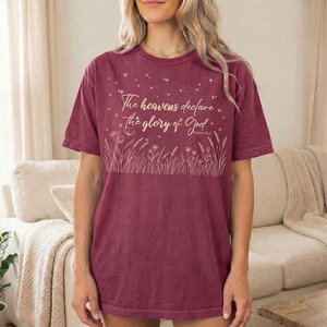 A woman wearing a navy blue t-shirt with a gold Psalm 19:1 night sky and botanical graphic, sitting indoors on a couch.