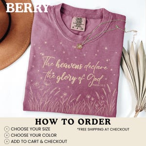 A berry-colored t-shirt with a gold starry botanical design, shown with a brown hat and dried grass on a white surface.