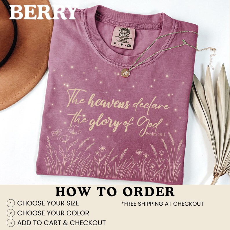 A berry-colored t-shirt with a gold starry botanical design, shown with a brown hat and dried grass on a white surface.