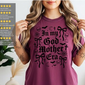 A person wearing a berry-colored t-shirt with the black In my God Mother Era gothic graphic.
