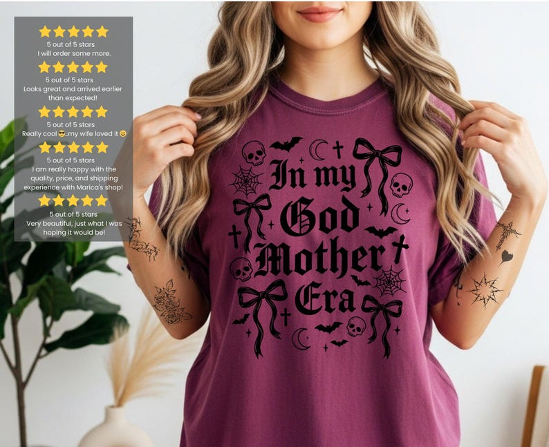A person wearing a berry-colored t-shirt with the black In my God Mother Era gothic graphic.