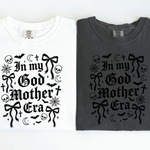 Two folded t-shirts side by side, one white and one dark gray, both printed with the black In my God Mother Era gothic design.