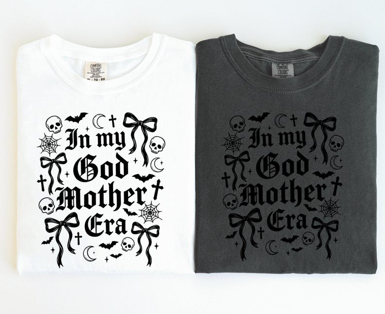 Two folded t-shirts side by side, one white and one dark gray, both printed with the black In my God Mother Era gothic design.