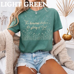 A person wearing a light green t-shirt with a gold Psalm 19:1 starry field design, denim shorts, and a beige cardigan.