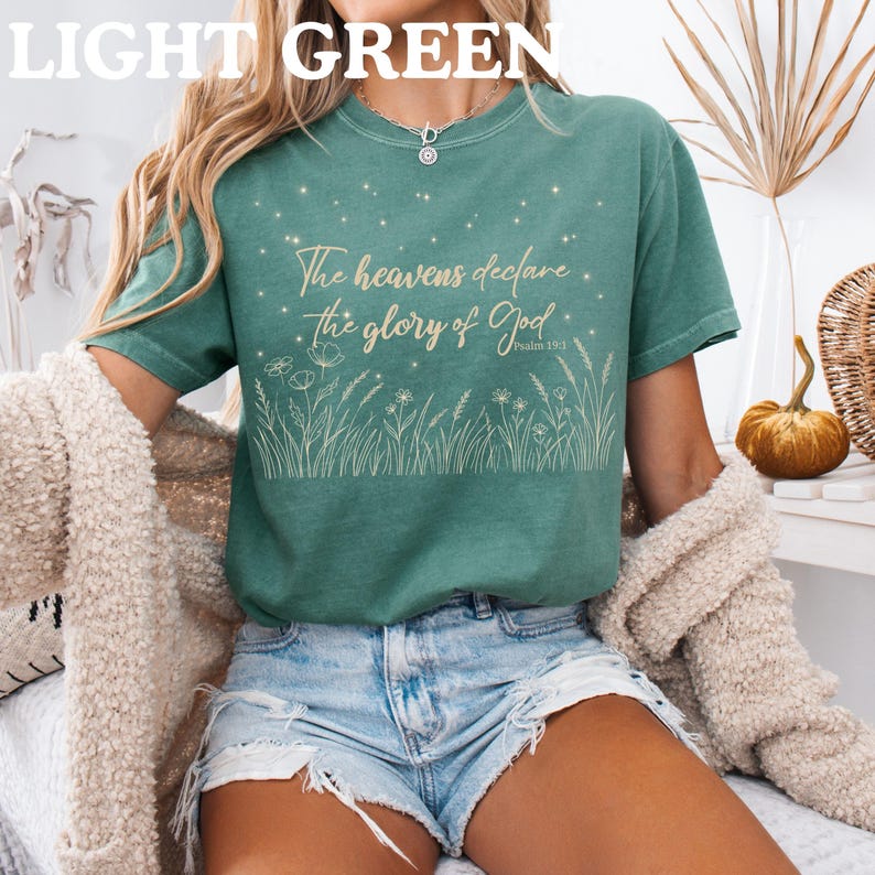 A person wearing a light green t-shirt with a gold Psalm 19:1 starry field design, denim shorts, and a beige cardigan.