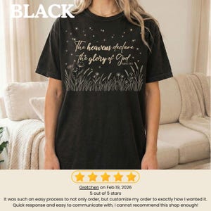 A person wearing a black crewneck t-shirt with a gold graphic of starry night sky above grass and the text The heavens declare the glory of God.