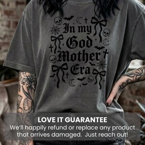 A person wearing a dark gray t-shirt with the same black In my God Mother Era gothic graphic design.