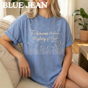 A woman wearing a blue jean colored t-shirt featuring a gold biblical star and wildflower graphic, sitting on a sofa.