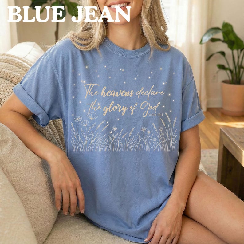 A woman wearing a blue jean colored t-shirt featuring a gold biblical star and wildflower graphic, sitting on a sofa.