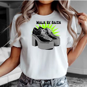 May include: White t-shirt with the text "WALK BY FAITH" above a graphic of black platform shoes with white laces. The shoes are set against a green starburst design. The text "2 Corinthians 5:7" is below the shoes.