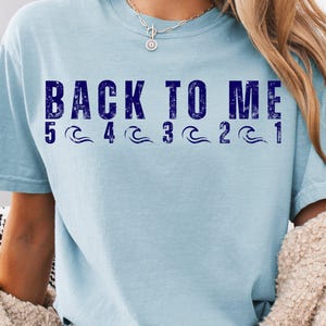 May include: Light blue t-shirt with the text "BACK TO ME" in dark blue block letters. Below the text are numbers 5 through 1 with wave graphics. The shirt has short sleeves and a crew neck.
