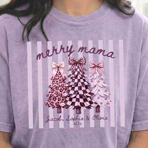 Könnte beinhalten: Ein hellviolettes T-Shirt mit einer Grafik von drei stilisierten Weihnachtsbäumen mit Schleifen. Die Bäume haben Leoparden-, Karomuster und Punkte. Der Text lautet merry mama und Jacob, Sophia & Olivia 2026 über vertikalen Streifen.